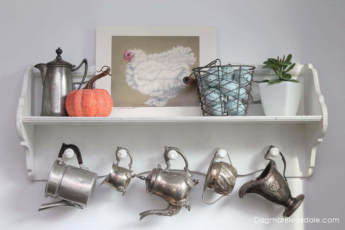 shelf with vintage decor, print of chicken, basket of eggs, pumpkin, plant, and silver creamers